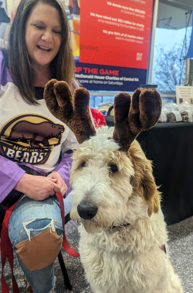 Maria Wright, BSN, RN sitting on a chair with the white and brown dog Cappuccino sitting in front of her. Cappuccino has fake reindeer antlers on his head.
